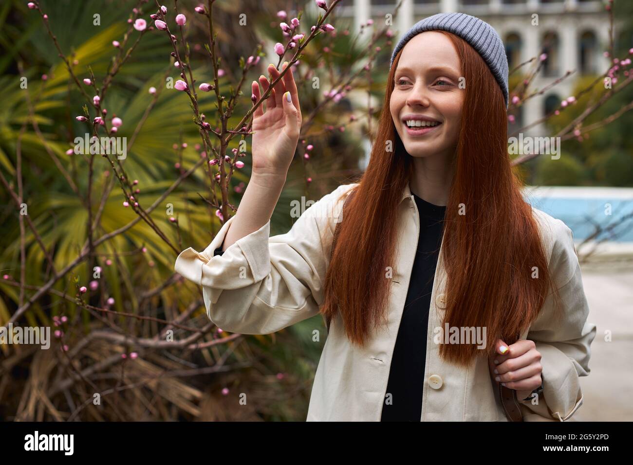 Sorprendente giovane donna dai capelli rossi in cappello che si pone in fiore albero a primavera. Bella giovane donna felice godendo l'odore nel giardino fiorente della molla, da sola, Foto Stock