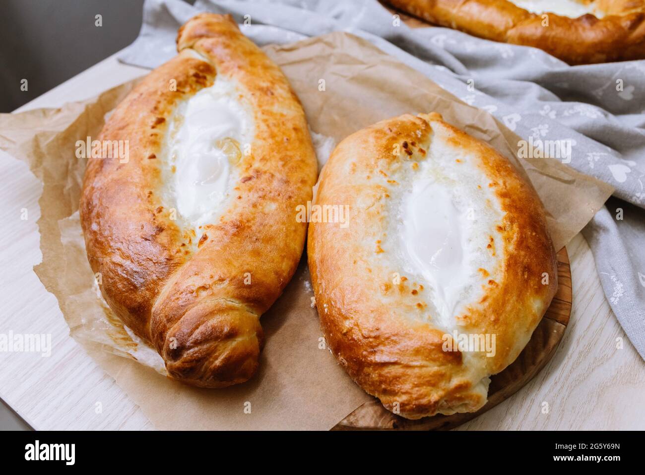 I khachapuri caldi in stile adjariano al forno con uova e formaggio fuso direttamente dal forno sono appoggiati su tavola di legno con un asciugamano grigio sulla destra Foto Stock