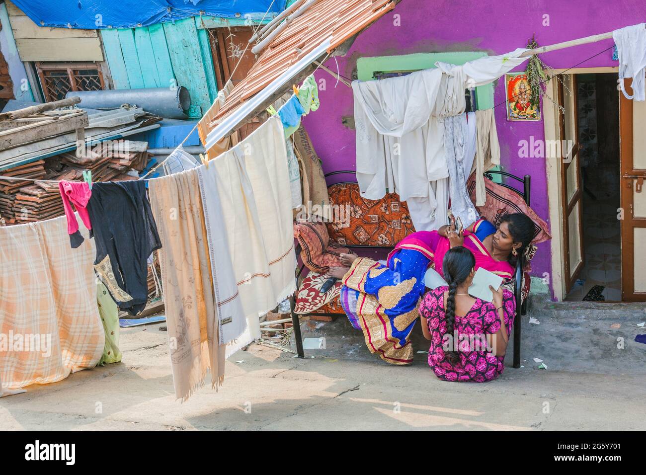 Due graziose femmine indiane che si rilassano al di fuori della loro colorata baracca con lenzuola appese dalla linea di vestiti, Trichy, Tamil Nadu, India Foto Stock