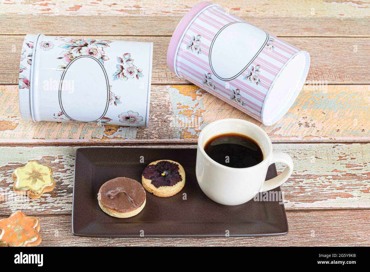 Biscotto alla burreria con un fiore secco sopra, accanto ad altri biscotti, tazza di caffè e lattine sdraiati. Foto Stock