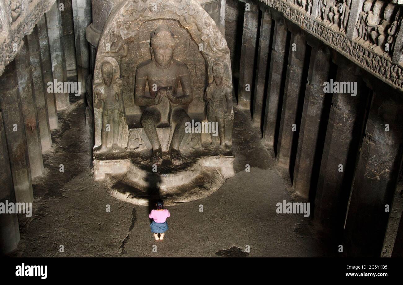 Una ragazza in un vestito rosso è seduta ai piedi del Buddha e pregando. C'è una statua molto grande e lineare di Buddha, grotte di Ellora, Aurangabad-in Foto Stock