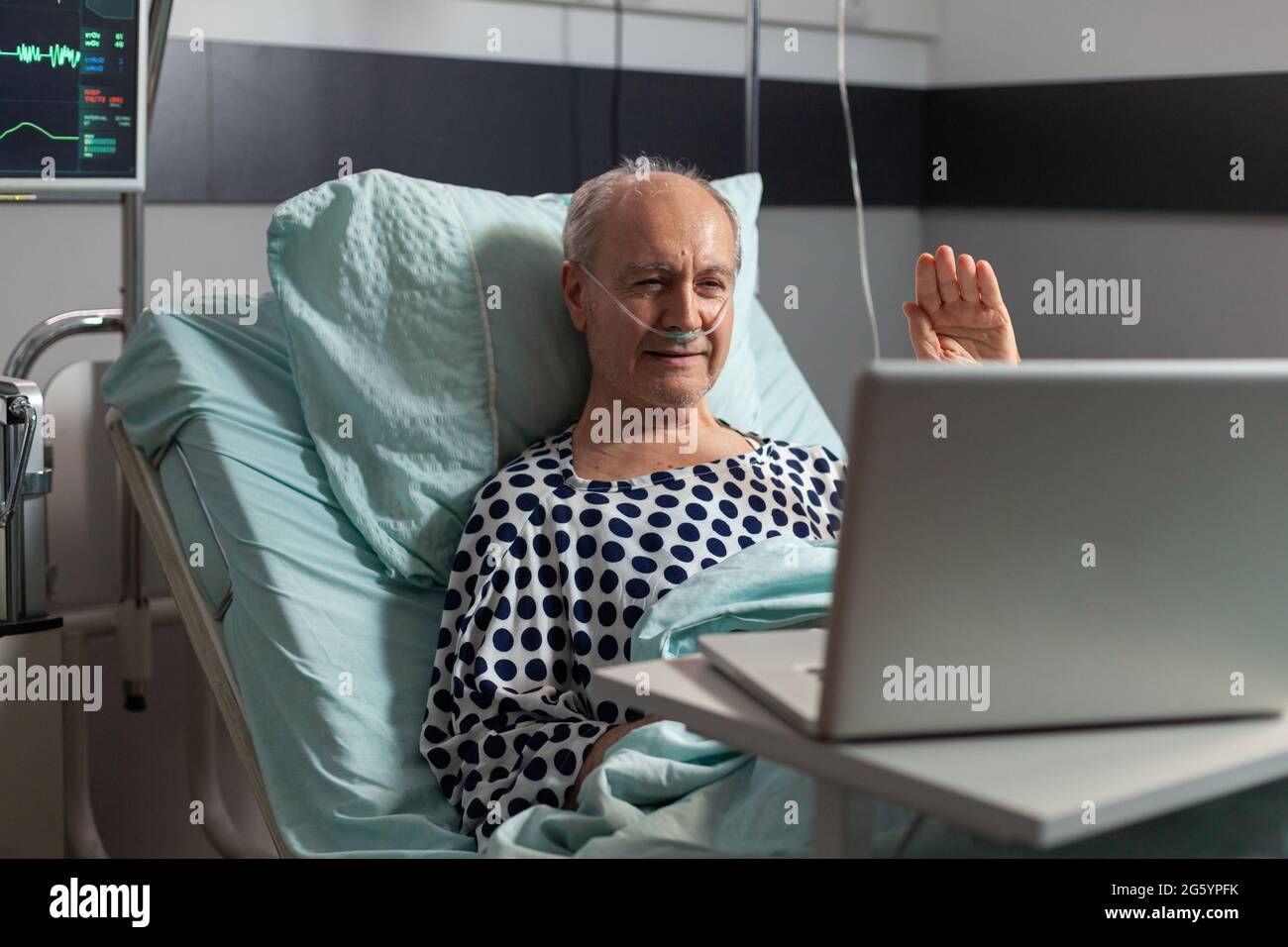 Ritratto dell'uomo anziano che saluta la famiglia a una macchina fotografica del portatile che si trova nel letto dell'ospedale, dopo la diagnosi della malattia, che respira con il tubo dell'ossigeno. Moderno eqipment per il monitoraggio della frequenza cardiaca paciente durante il recupero. Foto Stock