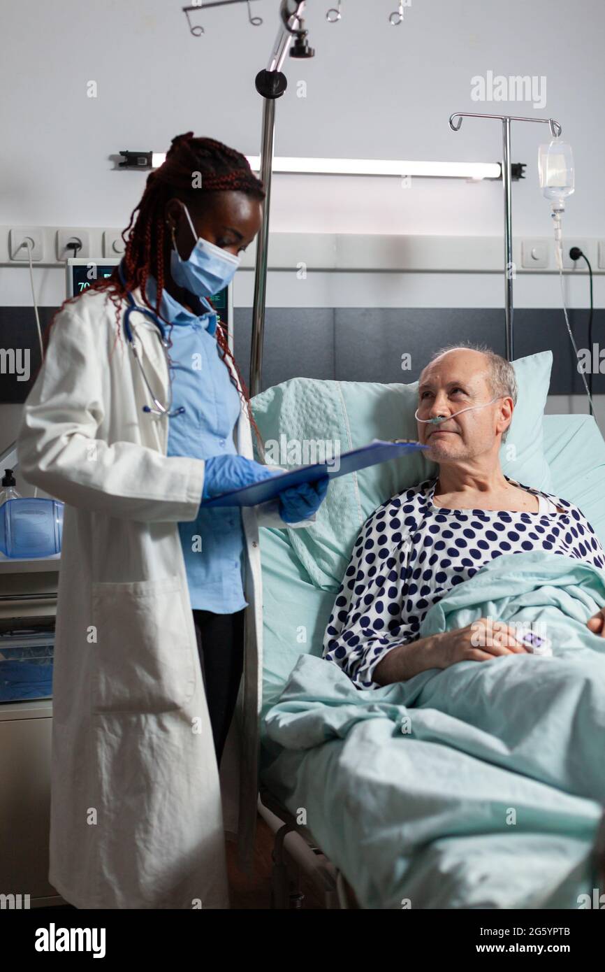 Medico afroamericano con maschera chirurgica che legge la diagnosi dalla clipboard a malato malato malato paziente anziano che si posa a letto, respirando con l'aiuto della maschera di ossigeno, ascoltando discutere con il personale medico circa il recupero. Foto Stock