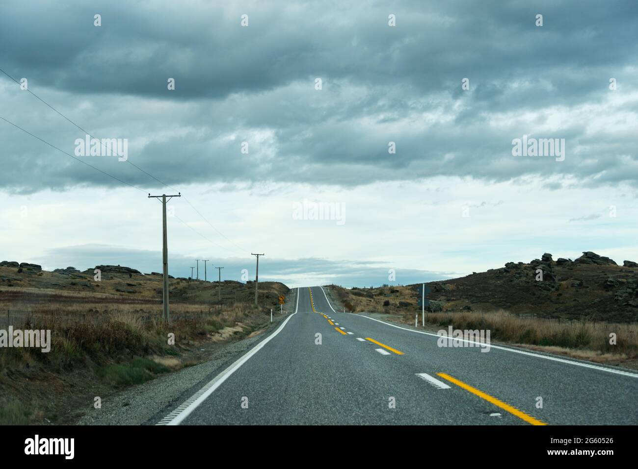 Una strada di campagna vuota con linee elettriche lungo la strada e nuvole scure in testa, la regione di Otago, Isola del Sud, Nuova Zelanda Foto Stock