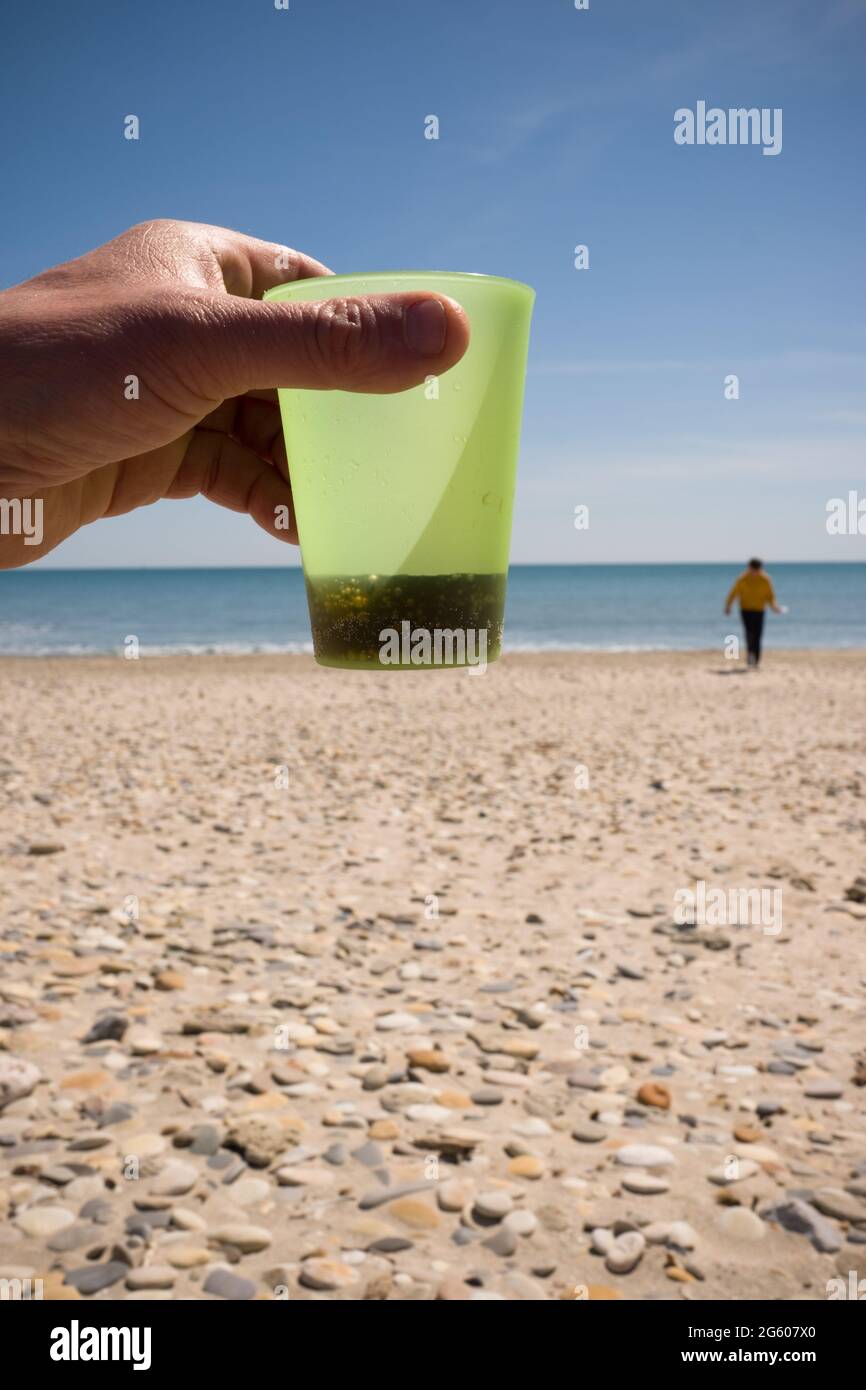 primo piano della mano che tiene in mano la tazza di plastica con coca cola con la persona sulla spiaggia dal mare sullo sfondo Foto Stock