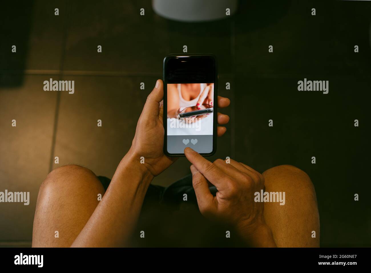 un giovane caucasico, seduto in bagno, risale online nel suo smartphone, con un'app simulata con la parola partita nel suo schermo Foto Stock