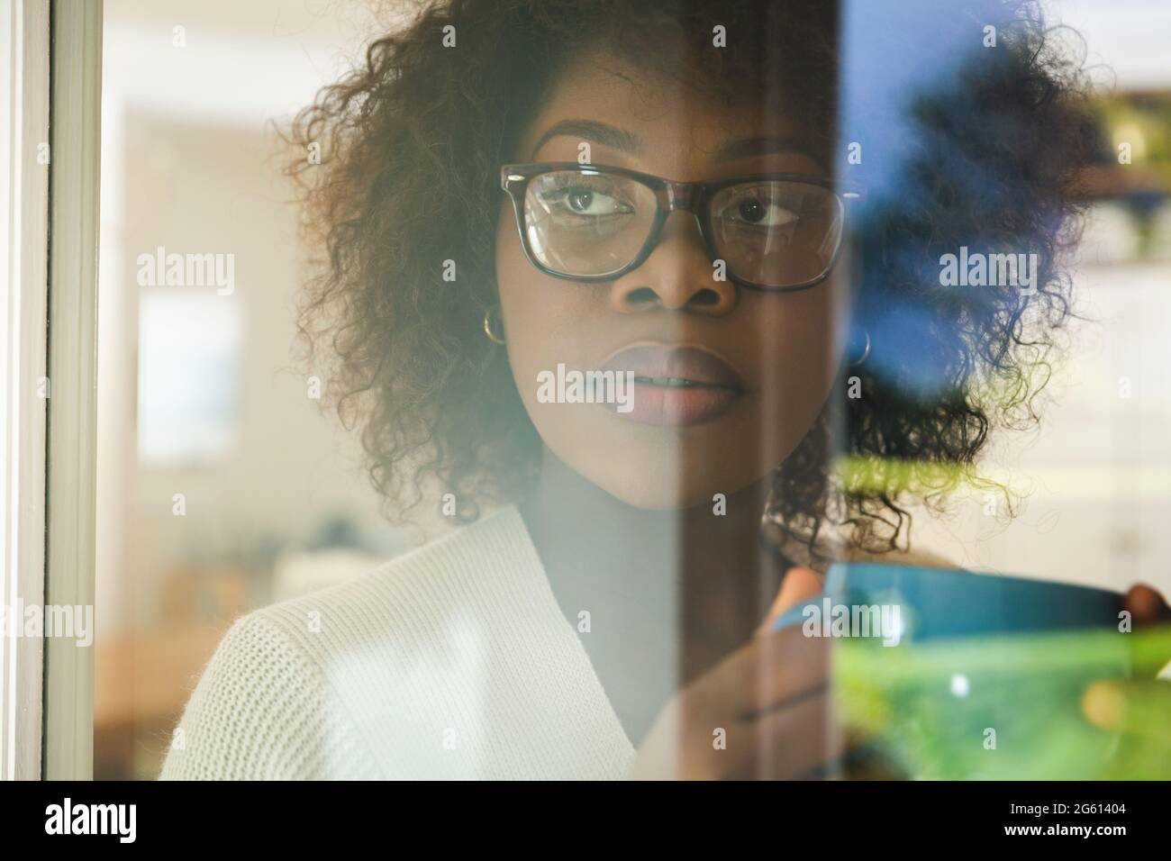 Donna afro-americana pensierosa che guarda fuori dalla finestra, tenendo una tazza di caffè Foto Stock