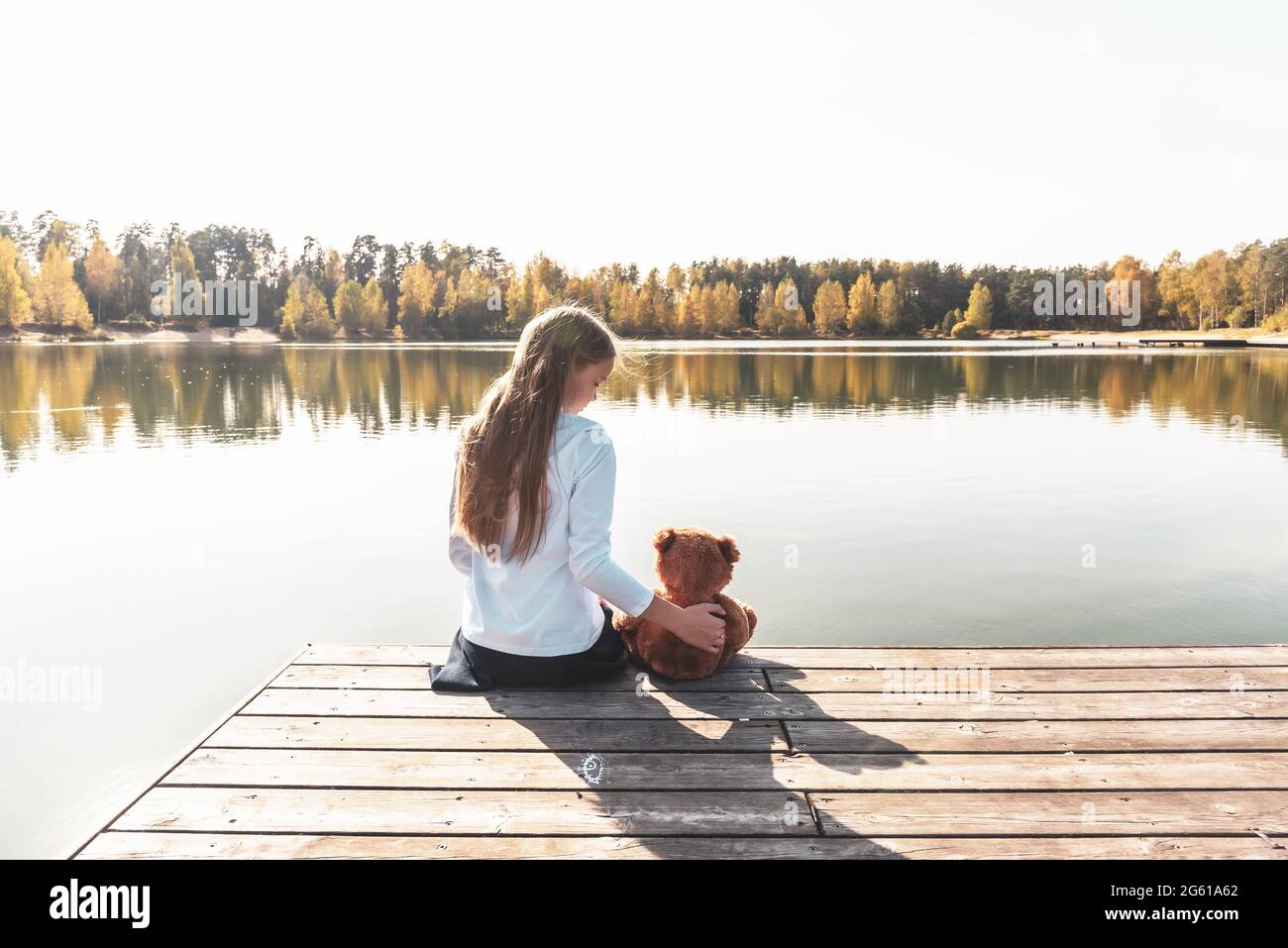 Ragazza con orsacchiotto siede sul molo di legno vicino a un ampio fiume Foto Stock