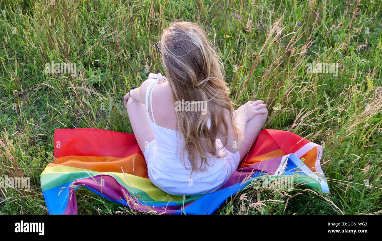 Donna bionda lesbica bisessuale seduta sulla bandiera LGBT sull'erba in abito bianco. Sostegno all'orientamento non tradizionale nel mese della dignità, giorno della bisessualità. Foto Stock