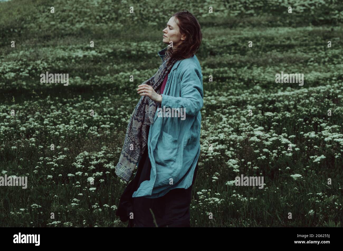 Ritratto laterale di una donna in cardigan di lino acqueo che cammina su un campo verde in una giornata ventosa. Concetto: Connessione con la natura, spiritualità, salute mentale Foto Stock