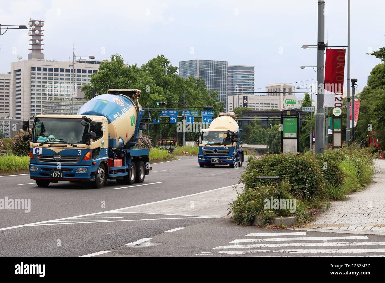 I camion per betoniere passano davanti ai cartelli olimpici di Tokyo appesi ai lampioni nell'area di Hayabusacho a Tokyo. Kasumigaseki è sullo sfondo. (6/2021) Foto Stock