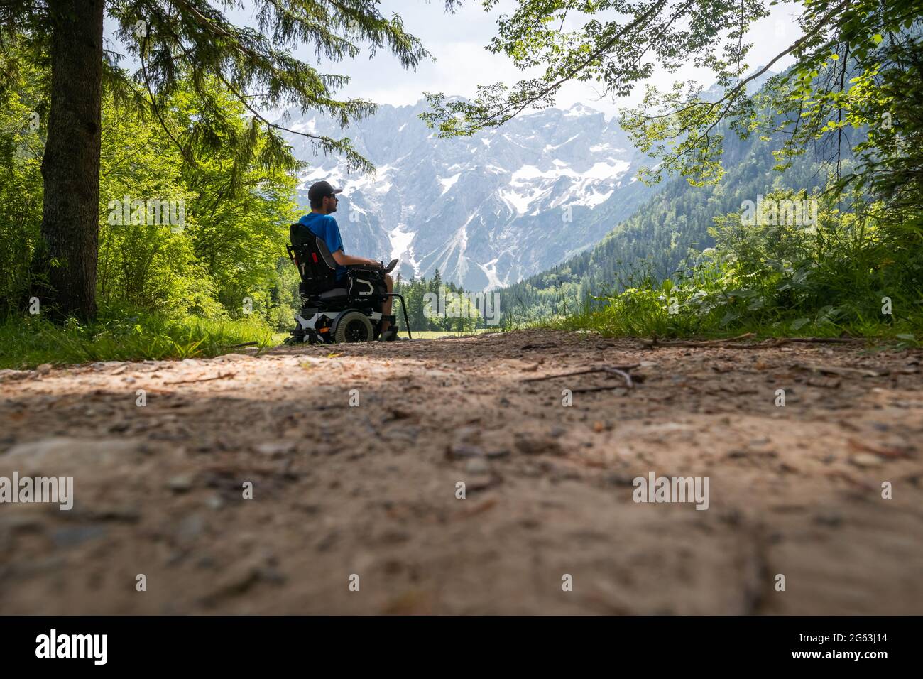 Uomo su una sedia a rotelle che si rilassa nella natura e guarda le montagne in una soleggiata giornata estiva. Foto Stock
