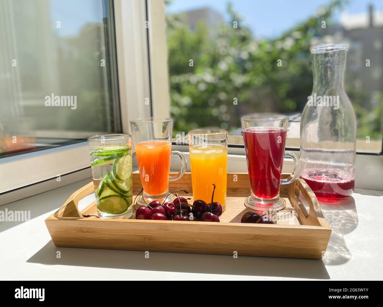 Vassoio di legno con bevande colorate, ciliegie su un davanzale. Decanter con i resti di succo rosso. Finestra aperta, estate calda, luce solare. Foto Stock