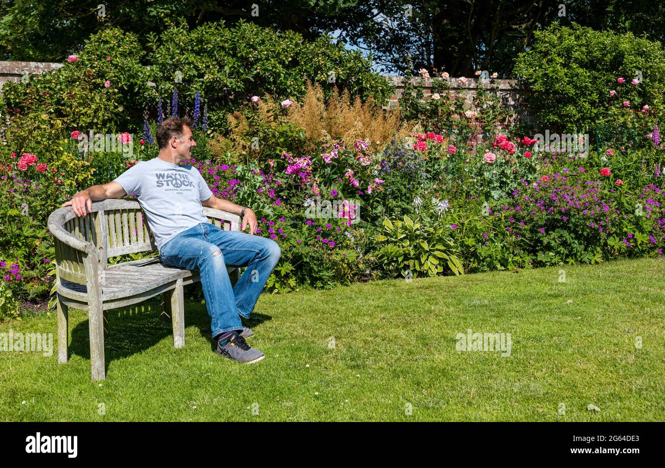 East Lothian, Scozia, Regno Unito, 2 luglio 2021. Scotland's Garden Scheme apertura di un nuovo giardino: Shane Corstorphine, ex CFO di Skyscanner e un appassionato giardiniere, ha trascorso 4 anni a preparare i giardini di Camptoun House per l'apertura per la prima volta per raccogliere fondi per la carità. Nella foto: Shane si rilassa su una panchina nel giardino murato con un colorato letto fiorito Foto Stock