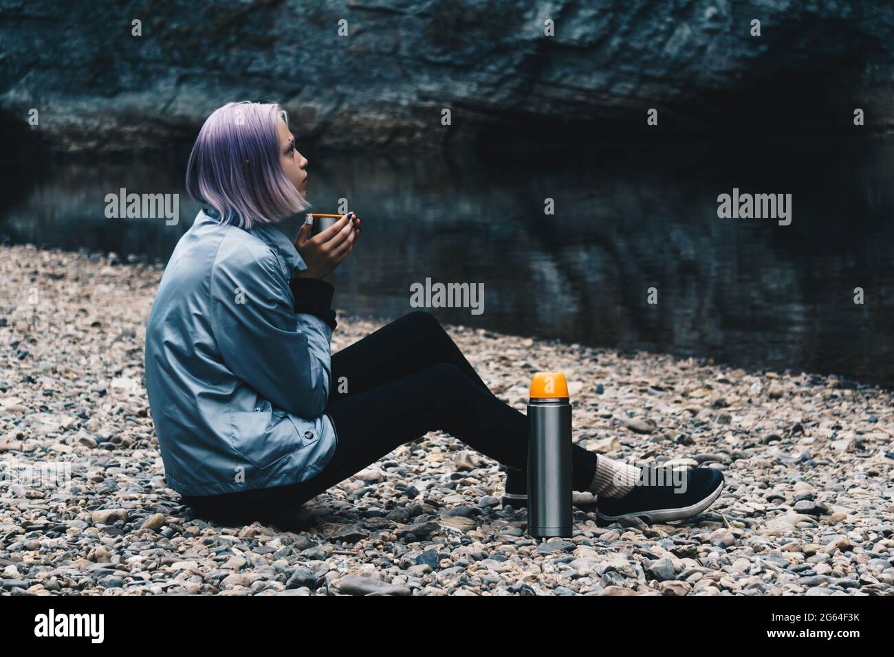 ragazza adolescente escursionista che riposa con una tazza sulla riva del fiume guardando le rocce costiere Foto Stock