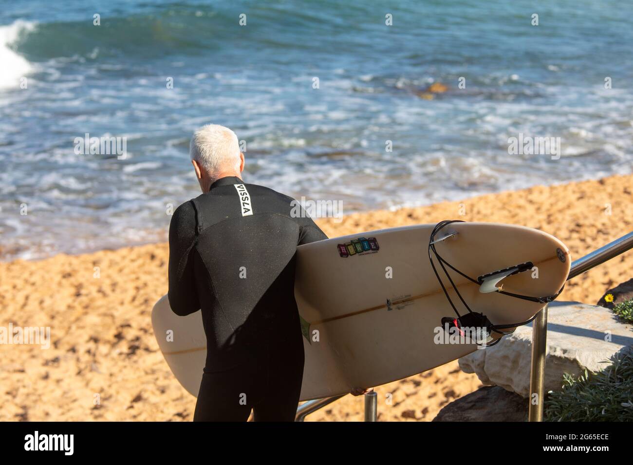 Uomo anziano con capelli grigi di media età che trasporta la tavola da surf verso l'oceano, Sydney, Australia Foto Stock