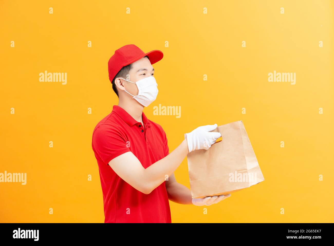 consegna guy hold takeaway pacchetto look fotocamera indossare maschera rosso t-shirt cappuccio isolato giallo colore sfondo Foto Stock