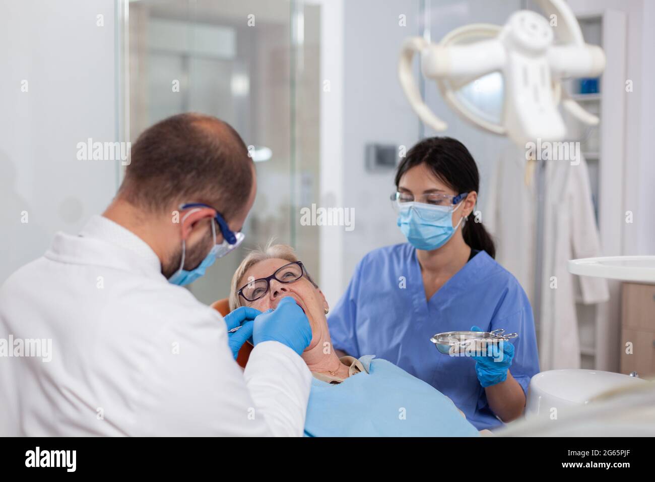 Stomatolog con assistente che mette il sigillo dentale sui denti della donna anziana. Paziente anziano durante la visita medica con dentista in studio dentistico con attrezzature arancioni. Foto Stock