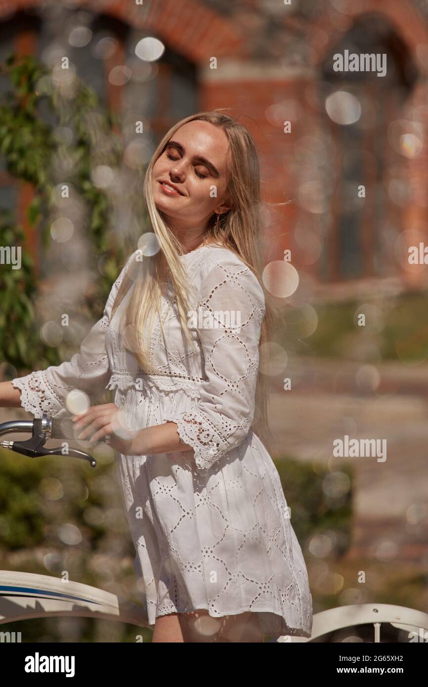 Bella ragazza bionda in abito bianco con la bici vicino alla fontana nella luce di sole Foto Stock