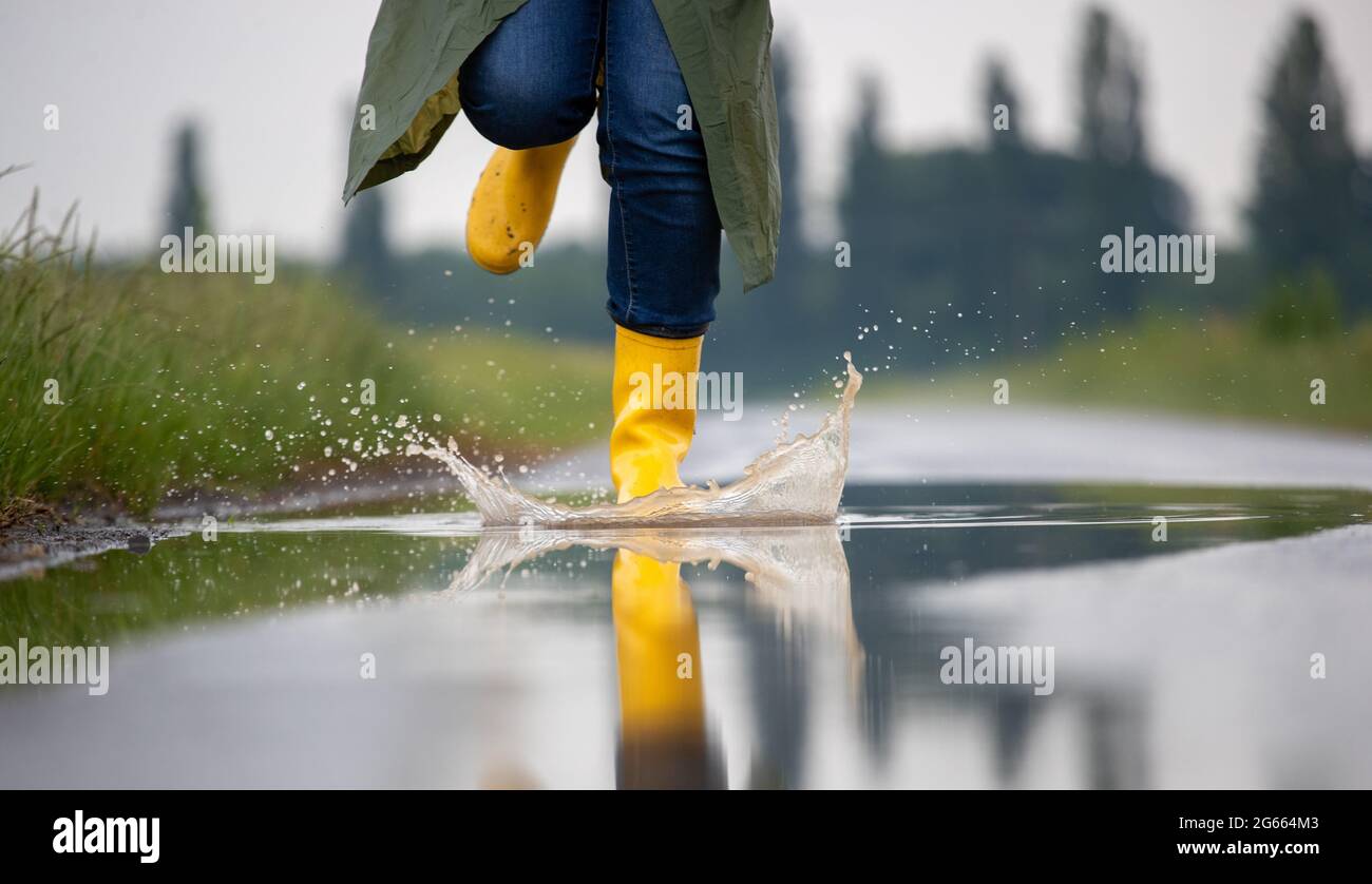 Primo piano delle gambe del contadino in stivali gialli e impermeabile verde che corrono su puddddles dopo la pioggia in campo Foto Stock