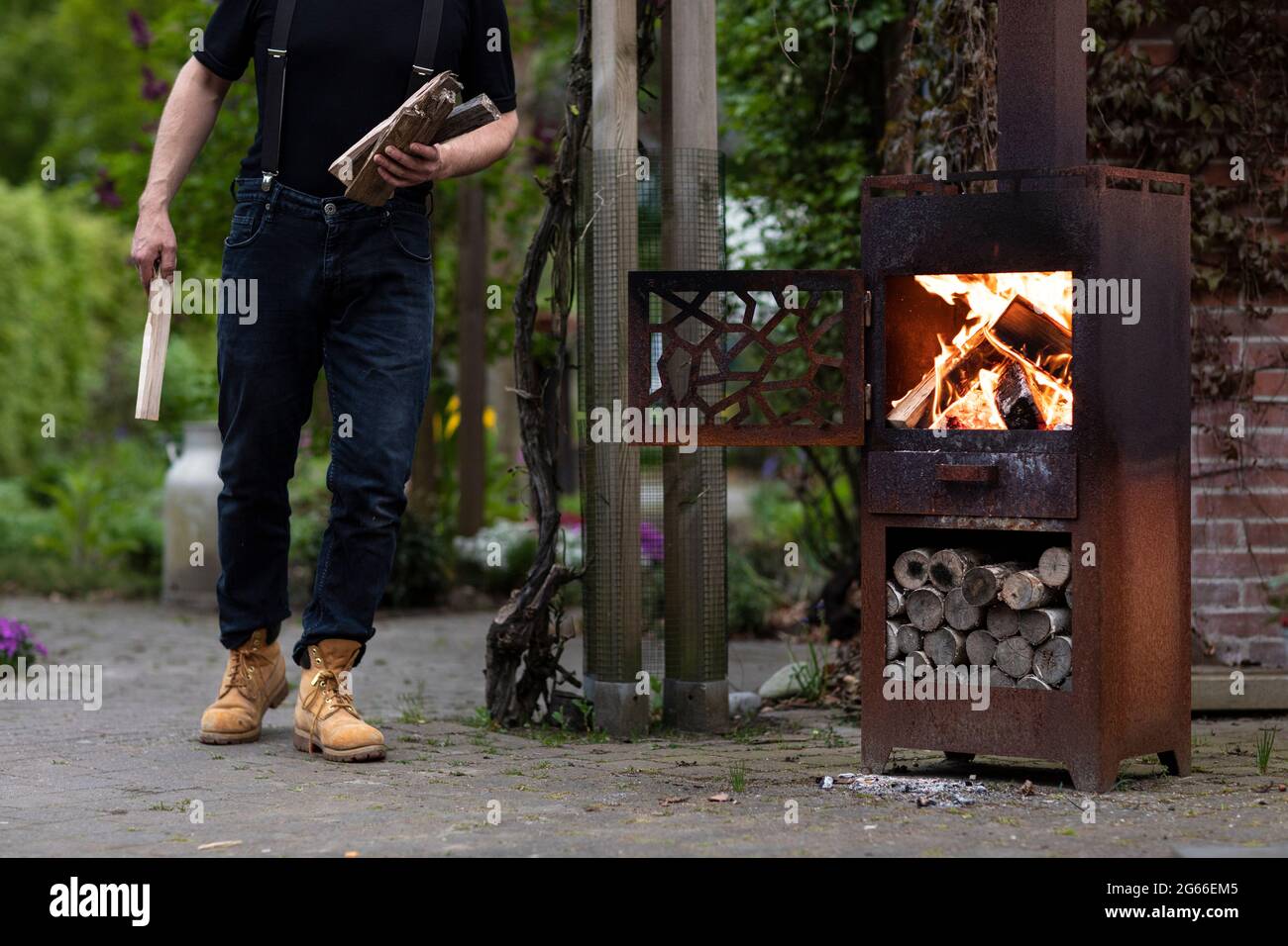 Un uomo che porta tronchi di legno ad una stufa a legna in metallo nel suo giardino mentre vive la vita semplice in una zona rurale con il verde Foto Stock