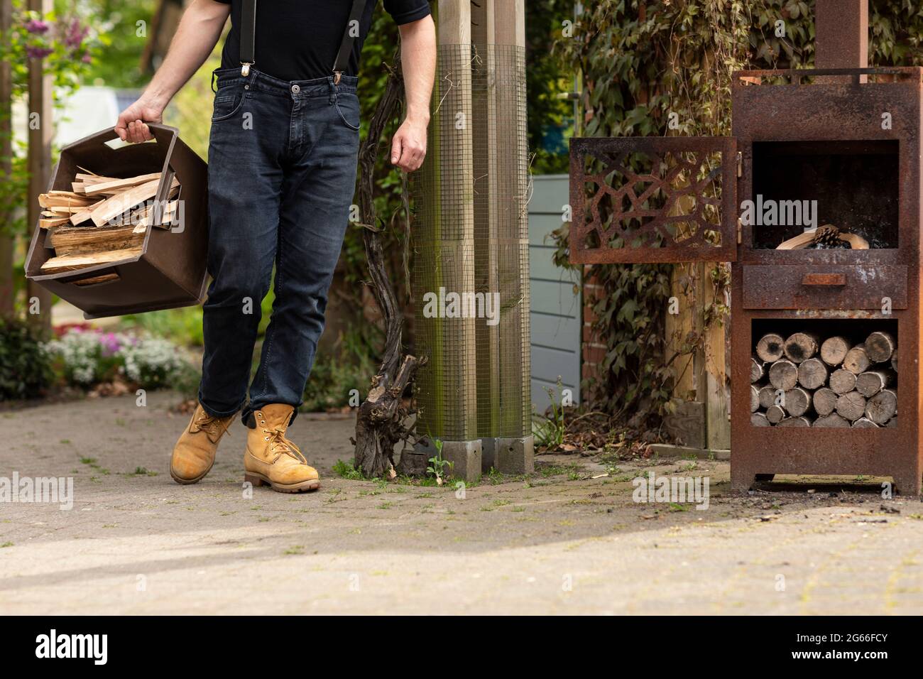 Uomo indossando abbigliamento casual che porta una grande scatola con tronchi di legno ad una stufa di legno di metallo nel suo giardino di cortile mentre vivono la vita semplice in un rurale A. Foto Stock