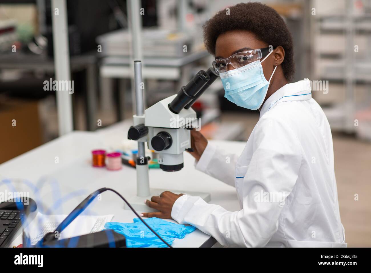Scienziato afroamericano donna in maschera e guanti di lavoro in laboratorio con microscopio. Ricerca e sviluppo di sostanze chimiche mediche da colore nero donna. Foto Stock