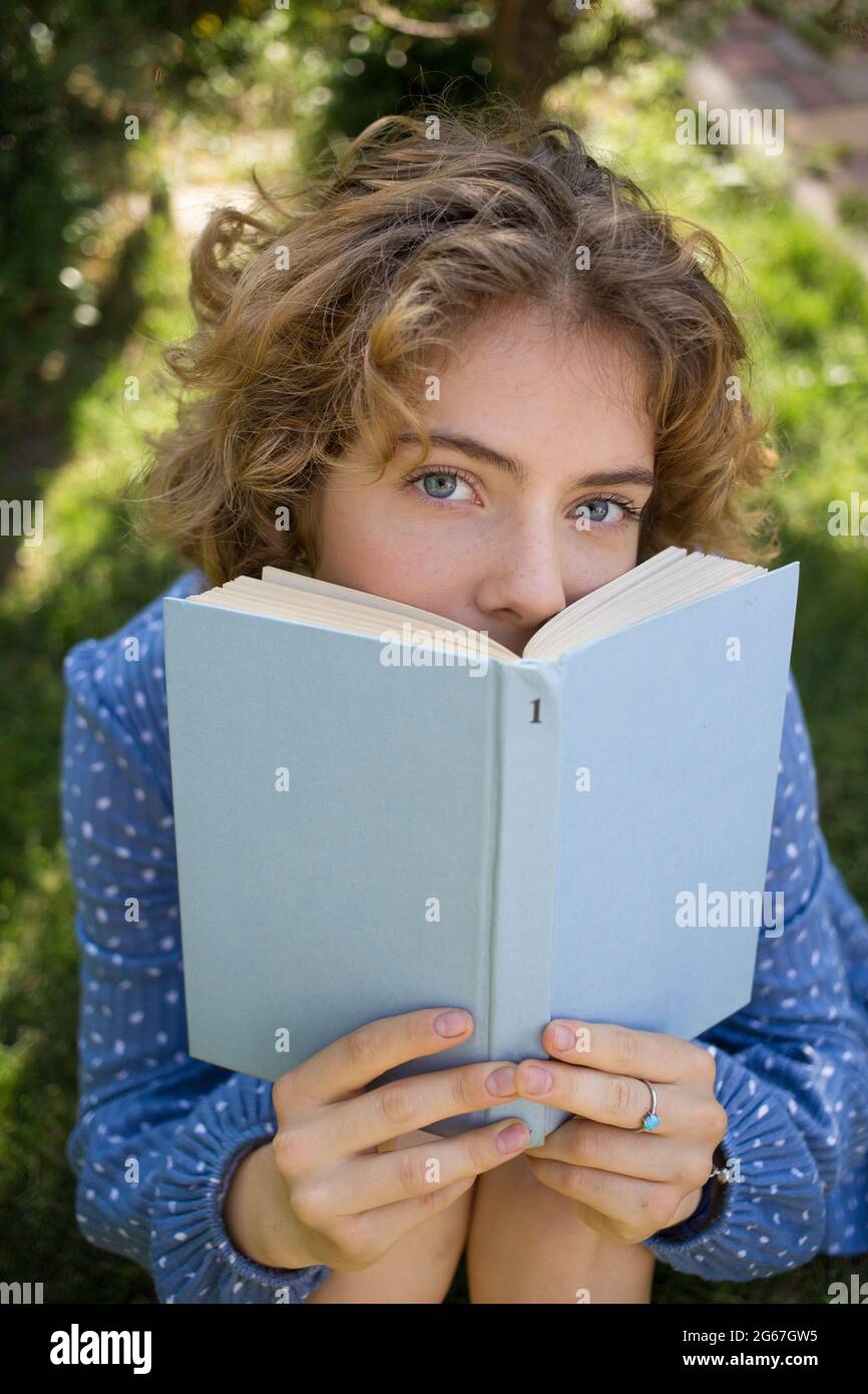 faccia di una ragazza carina - un adolescente 17 - 18 anni è nascosto dietro un libro. Occhi brillanti e belli. Preparazione di uno studente per gli esami. Tempo libero estivo. Passi Foto Stock