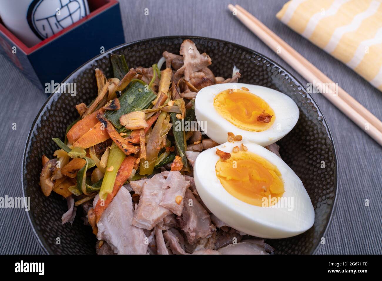 Studio di ciotola nera di tagliatelle di ramen con uova bollite, verdure e tacchino si incontrano, pronti per essere versati con brodo bollente. Bastoncini di legno Foto Stock