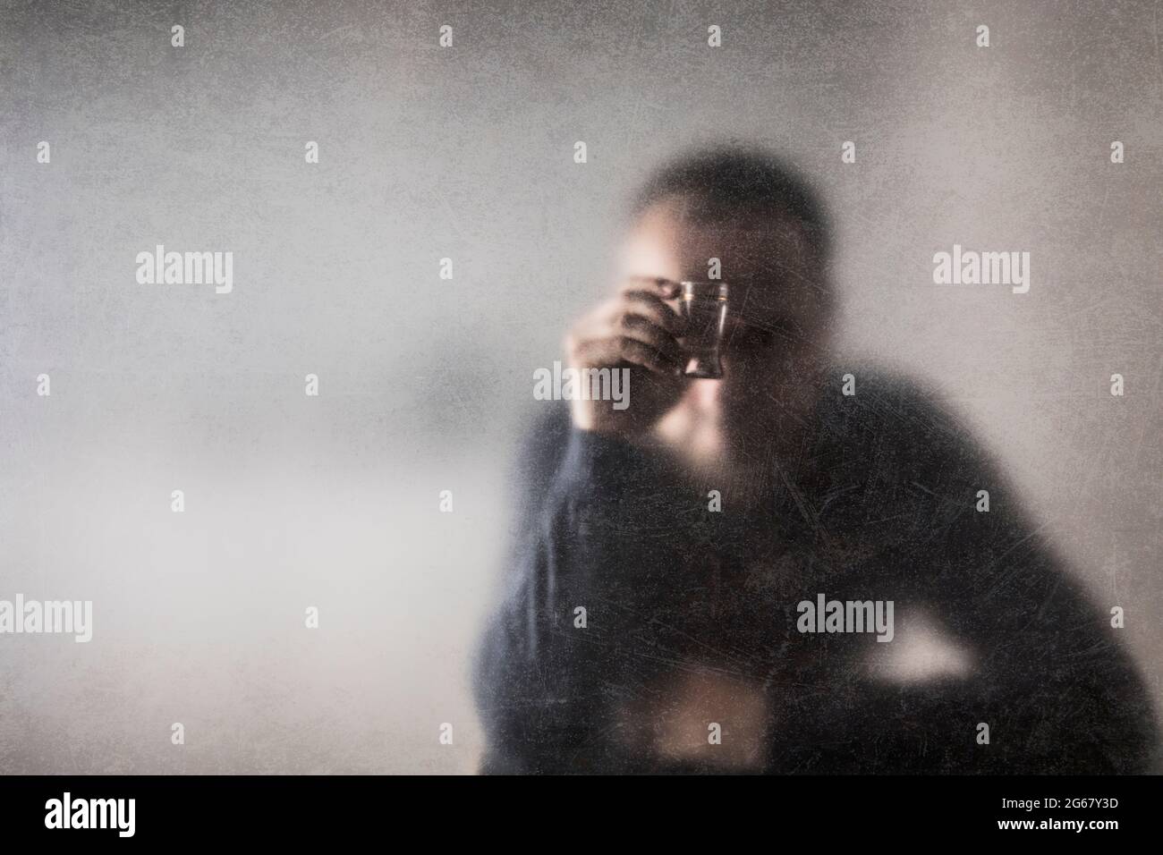 Uomo con un bicchiere di vodka dietro un vetro polveroso graffiato. Concetto di alcolismo. Foto Stock