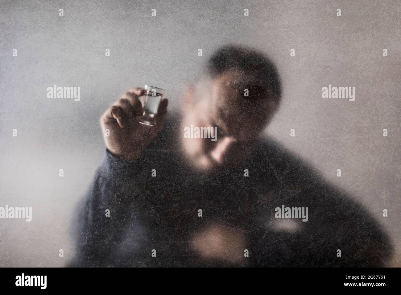 Uomo con un bicchiere di vodka dietro un vetro polveroso graffiato. Concetto di alcolismo. Foto Stock
