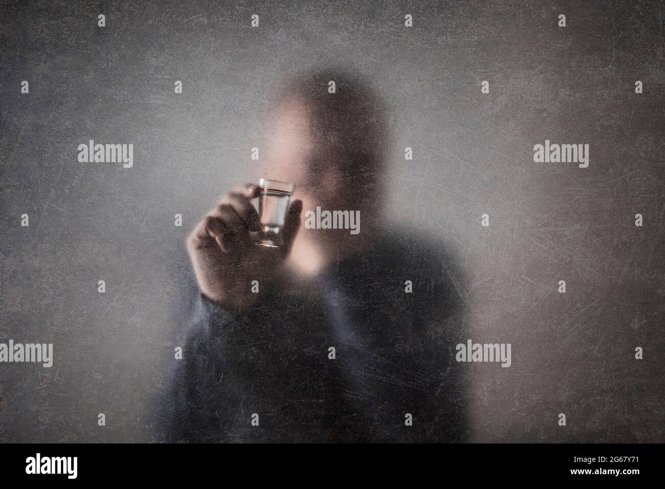 Uomo con un bicchiere di vodka dietro un vetro polveroso graffiato. Concetto di alcolismo. Foto Stock