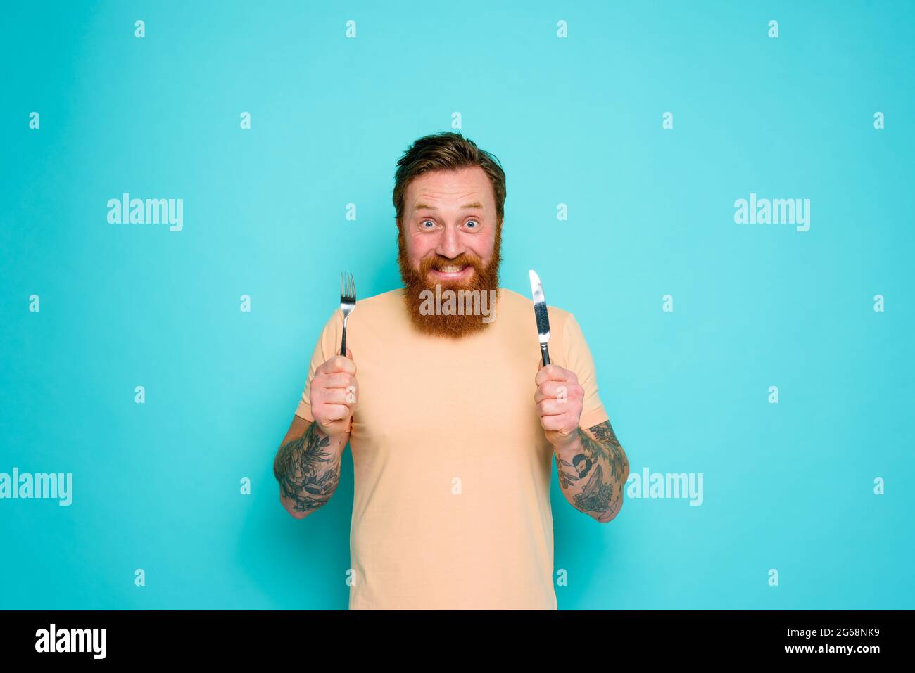 L'uomo felice con i tatuaggi è pronto a mangiare con le posate in mano Foto Stock