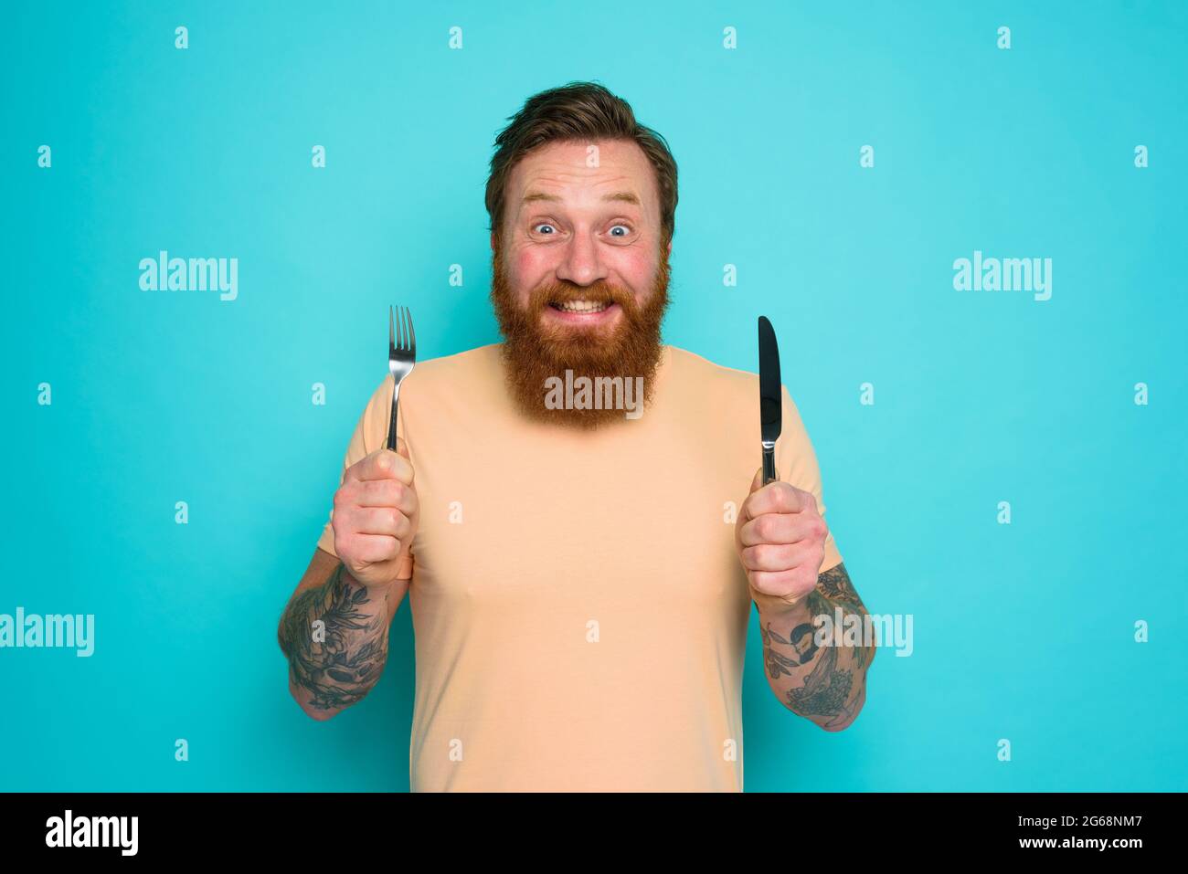 L'uomo felice con i tatuaggi è pronto a mangiare con le posate in mano Foto Stock