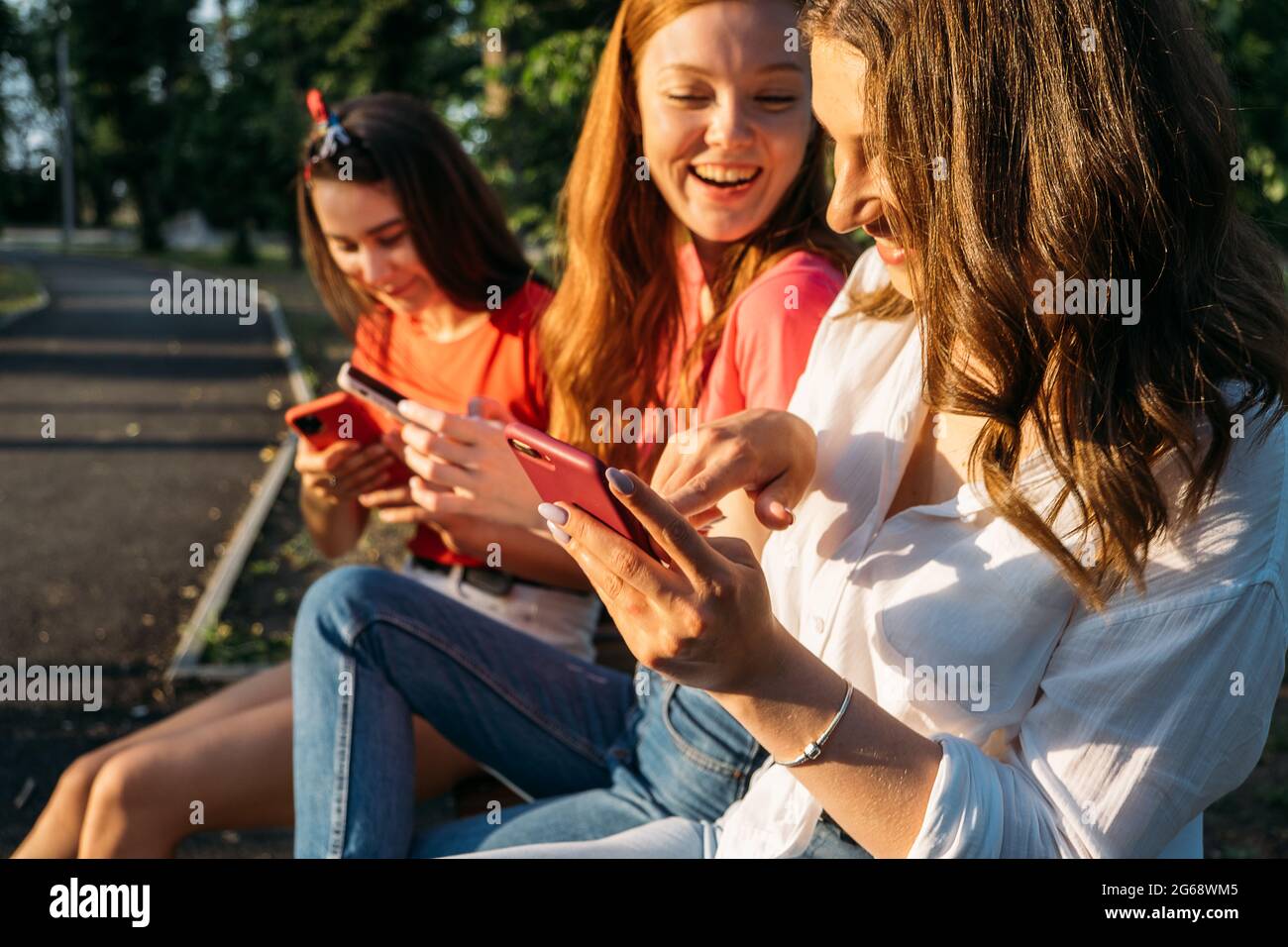 Tre amiche che guardano telefoni cellulari intelligenti e si divertono  seduti sulla panchina nel parco pubblico. Gen z, dipendenza dei dispositivi  degli adolescenti, detox digitale Foto stock - Alamy