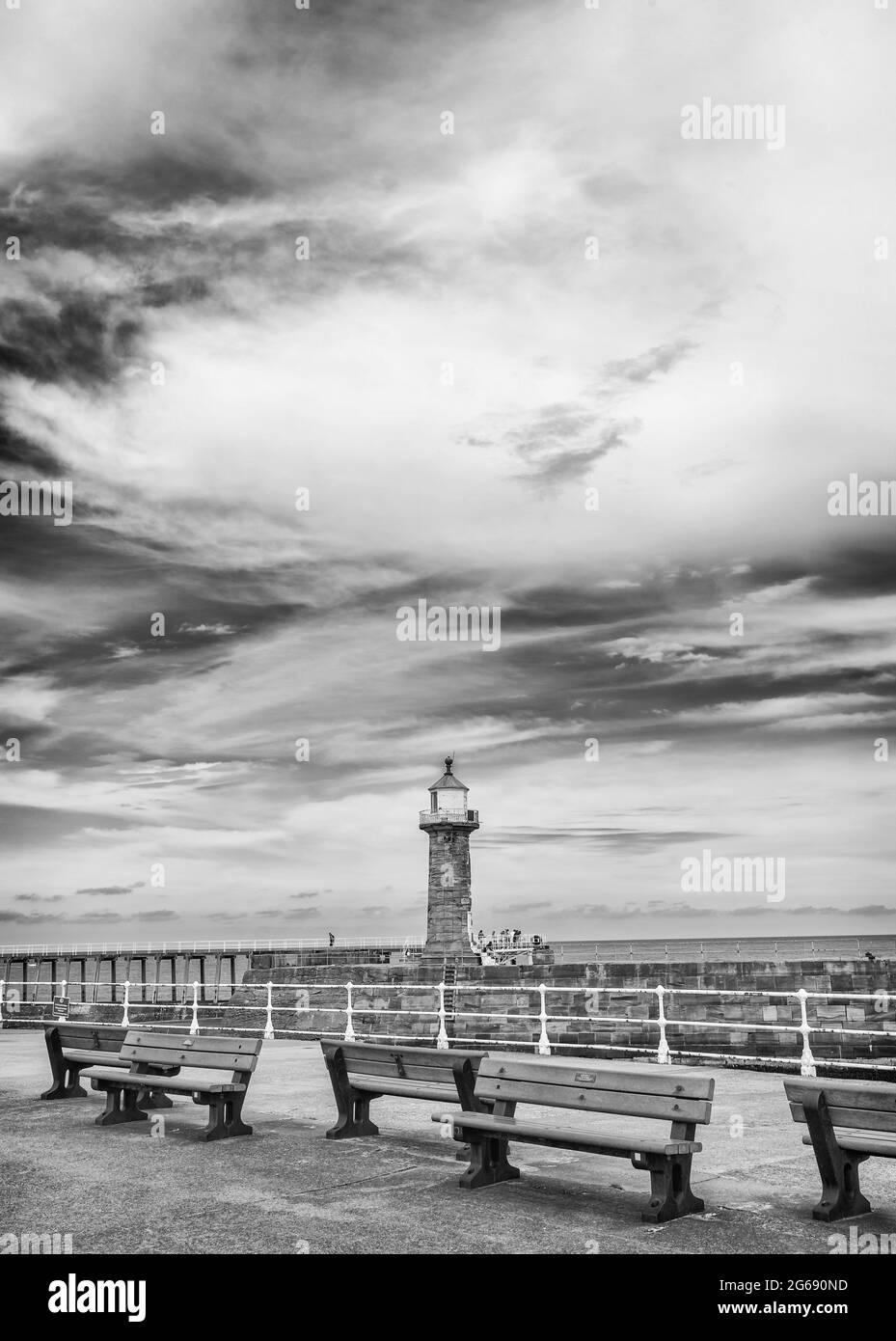 Un molo con panchine è in primo piano e oltre è un altro molo con un faro. Un cielo con nuvole è sopra. Foto Stock