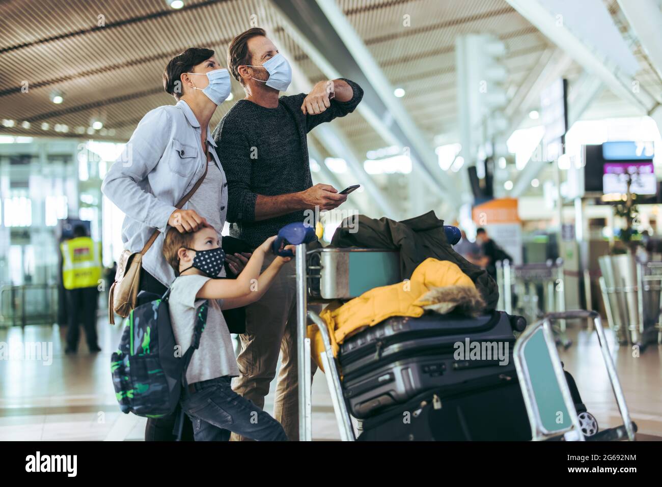 Famiglia in maschere di faccia che guardano i loro orari di volo a bordo all'aeroporto. Uomo turistico con moglie e bambino in attesa all'aeroporto con bagagli sul trolley durin Foto Stock
