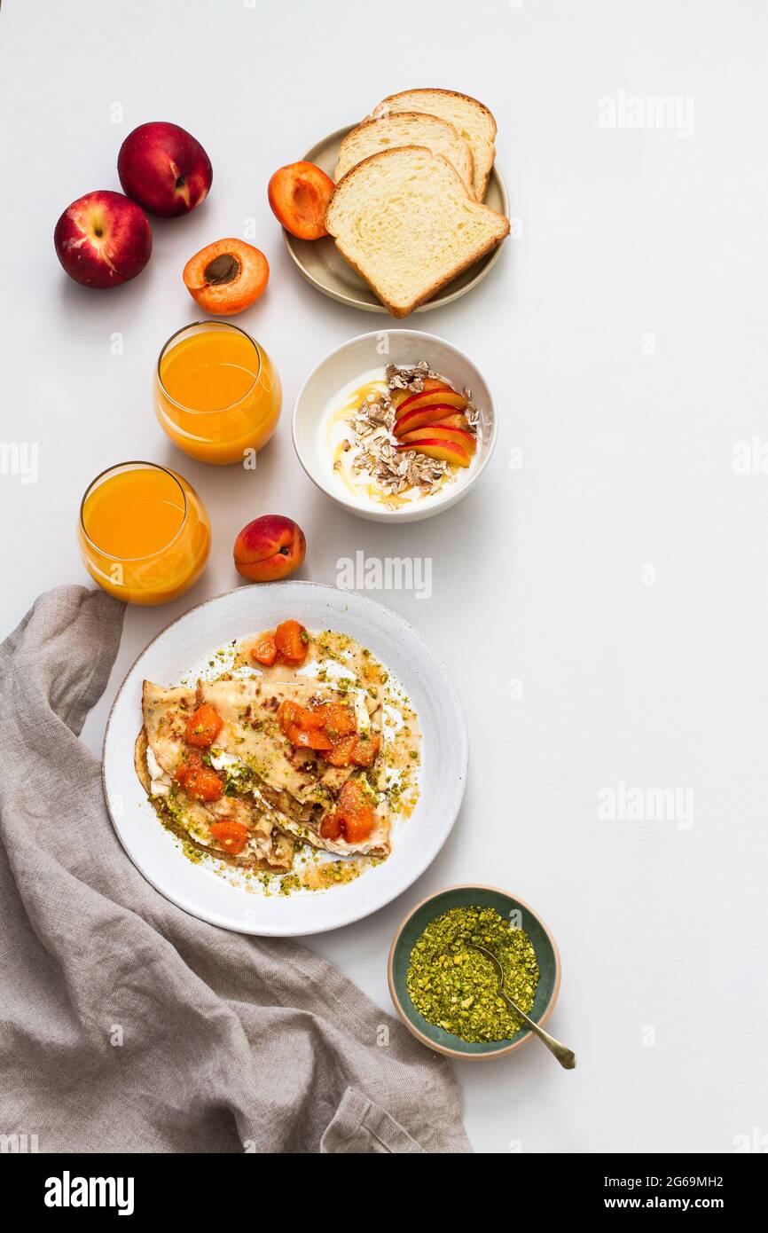 Colazione estiva con brioche, succo di frutta, albicocca e nettarine, yogurt con muesli, crepes con composta di albicocche e pistacchio. Foto Stock