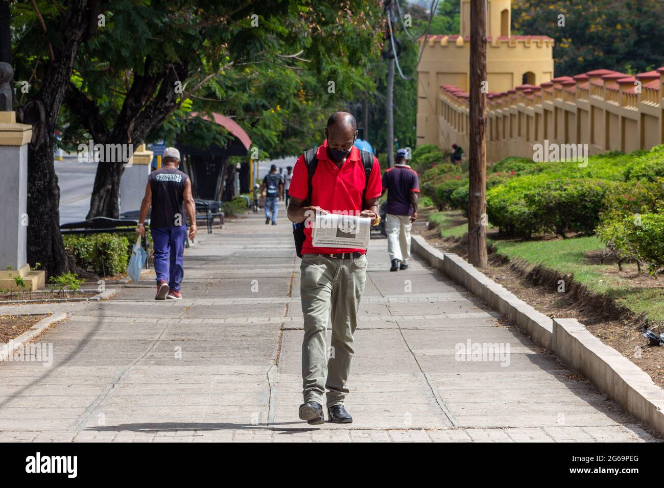 Un vero cubano di etnia afro-caraibica legge un giornale mentre cammina vicino alle mura di Cuartel Moncada a Santiago de Cuba, Cuba. Indossa un professionista Foto Stock