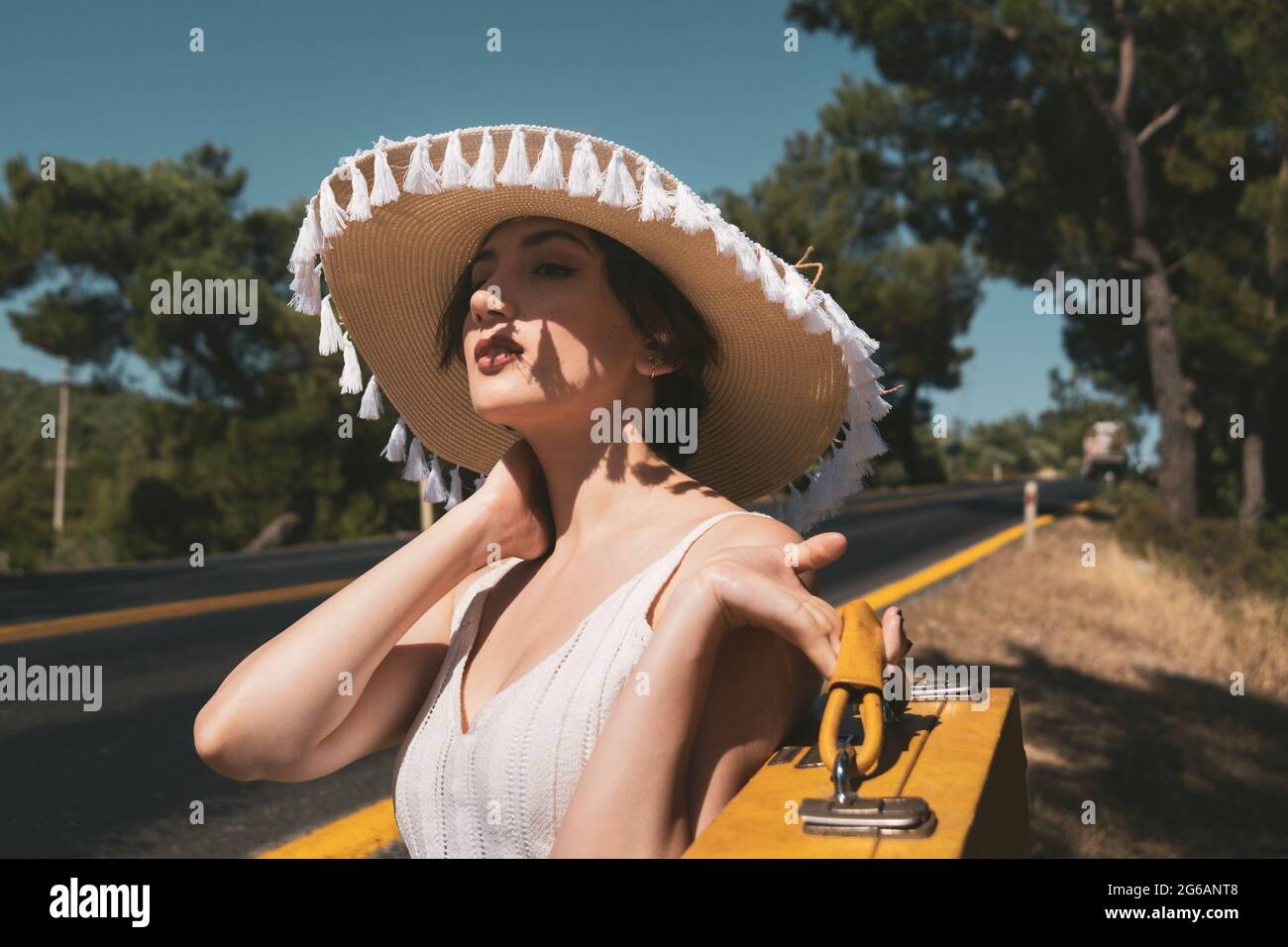 20s età una donna con un cappello di paglia, Jean shorts, maglia t-shirt e valigia gialla hitchhiking vicino alla strada paese Foto Stock