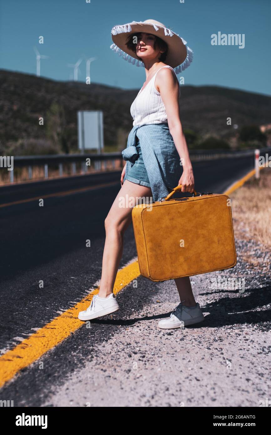 20s età una donna con un cappello di paglia, Jean shorts, maglia t-shirt e valigia gialla hitchhiking vicino alla strada paese Foto Stock