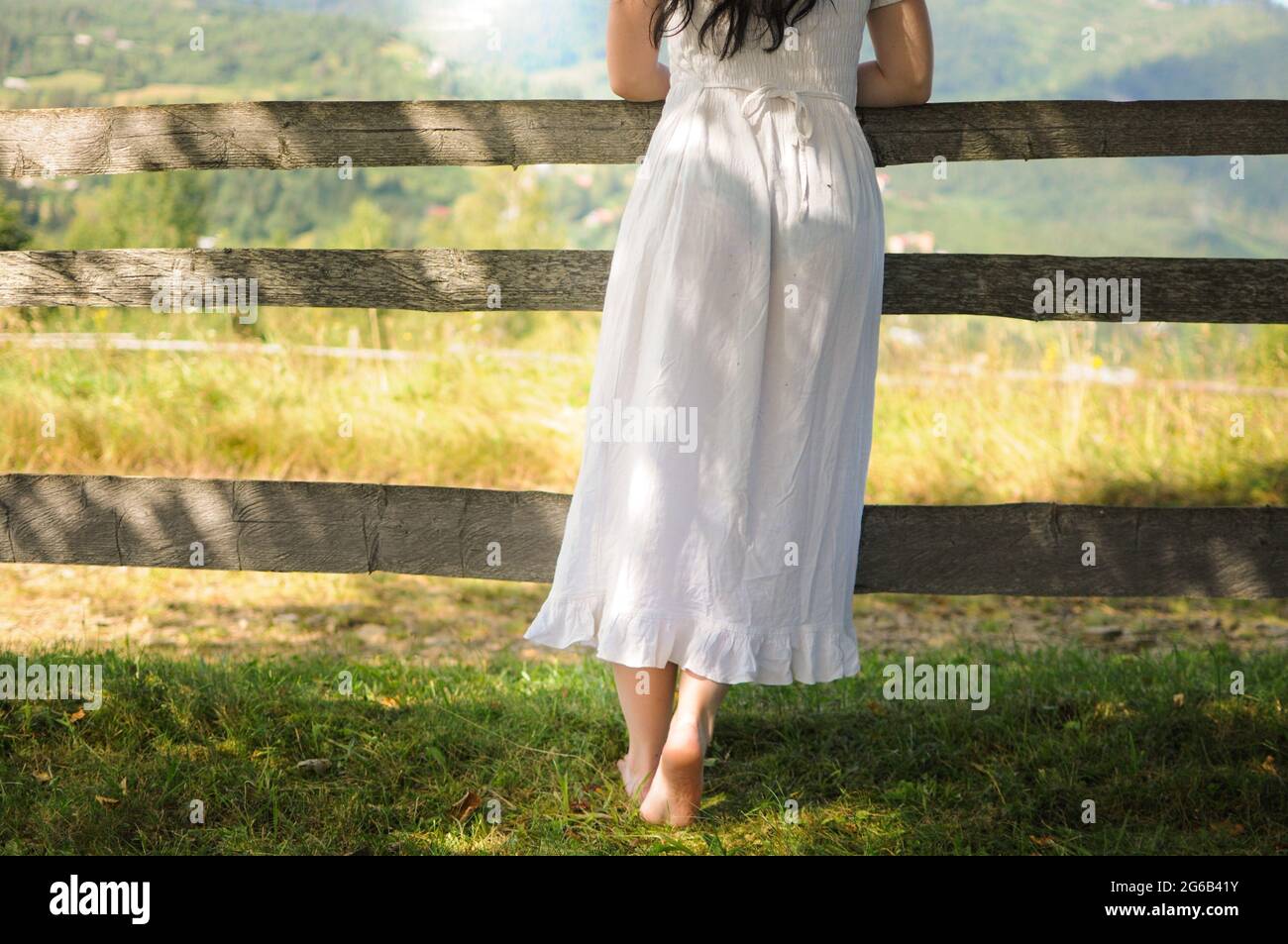 Una donna in abito bianco sta in piedi vicino a una recinzione di legno. In stile naturale in estate Foto Stock