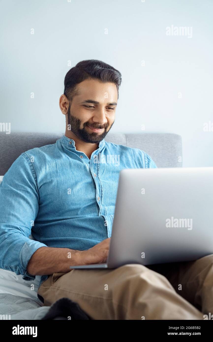 Sorridente uomo d'affari indiano che ha una chat informale di videochiamata sul laptop seduto a letto Foto Stock
