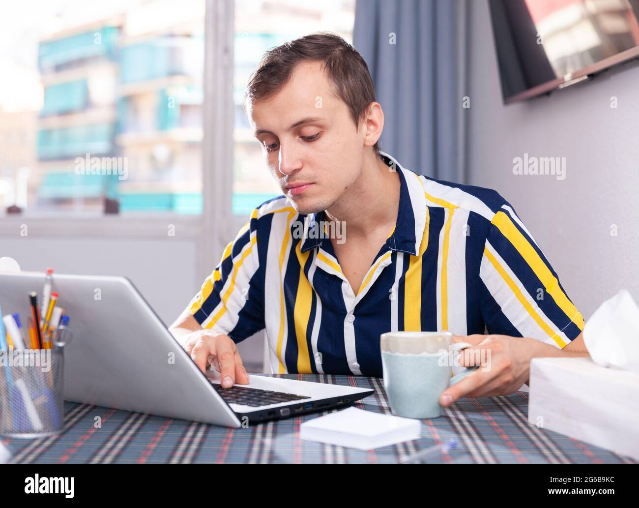 Il giovane si siede di fronte a un computer portatile aperto e beve il tè Foto Stock