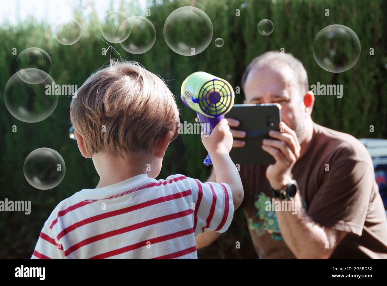 Uomo che fotografa suo figlio giocando con una pistola a bolle di sapone in giardino Foto Stock