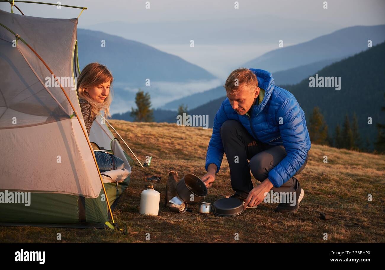 Campeggio in montagna. Uomo escursionista seduto squattando e versando acqua per fare caffè vicino a sua moglie, che ammirano la bellezza circostante all'alba dalla loro tenda al mattino. Foto Stock