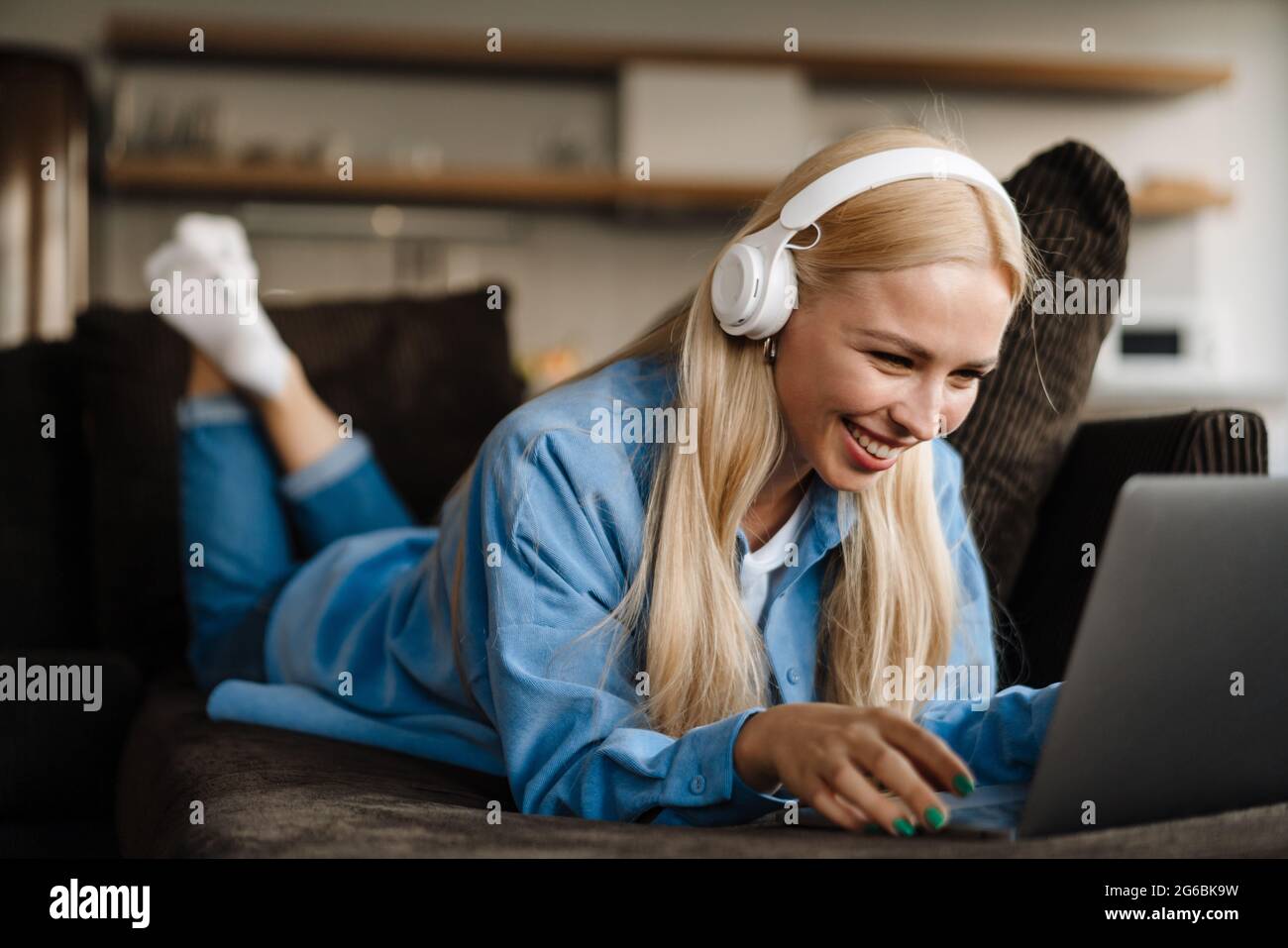 Donna bionda bianca in cuffia che usa il computer portatile mentre si sdraia sul divano a casa Foto Stock