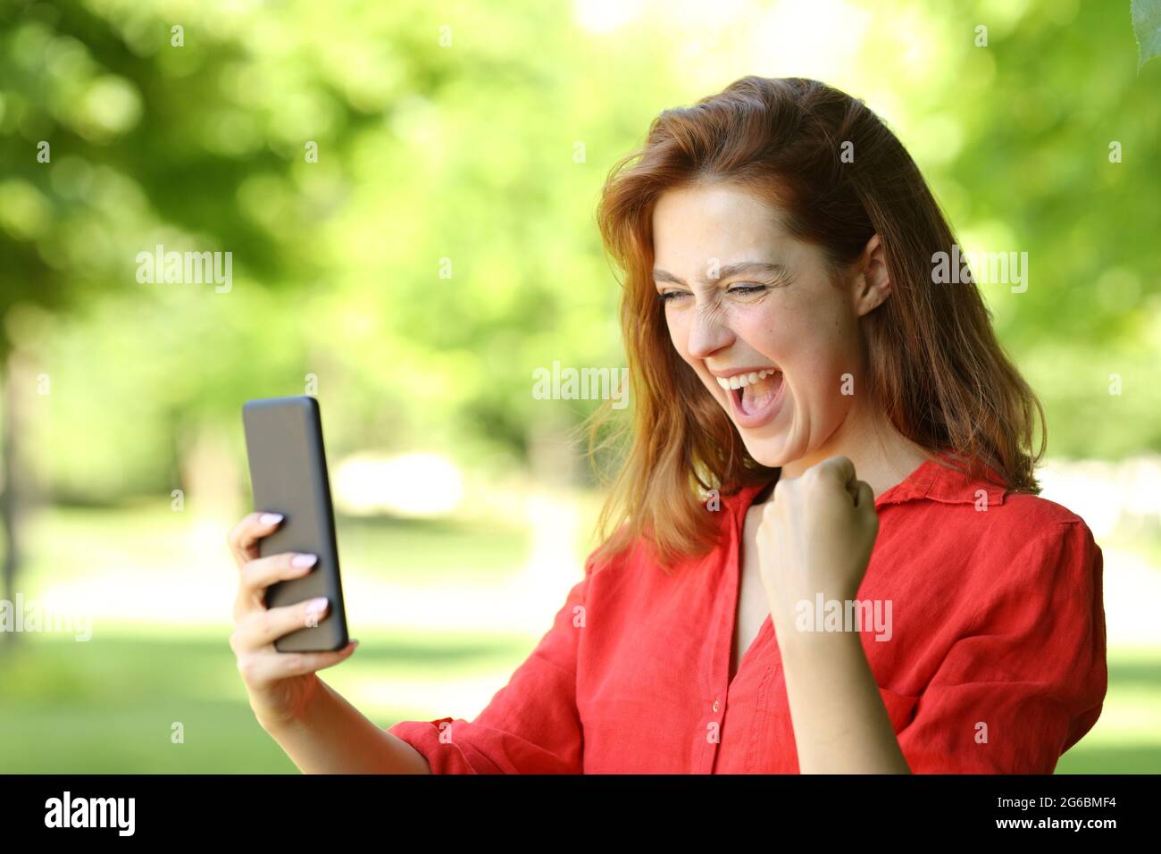 Donna entusiasta in rosso che festeggia le buone notizie controllando lo smartphone in un parco Foto Stock