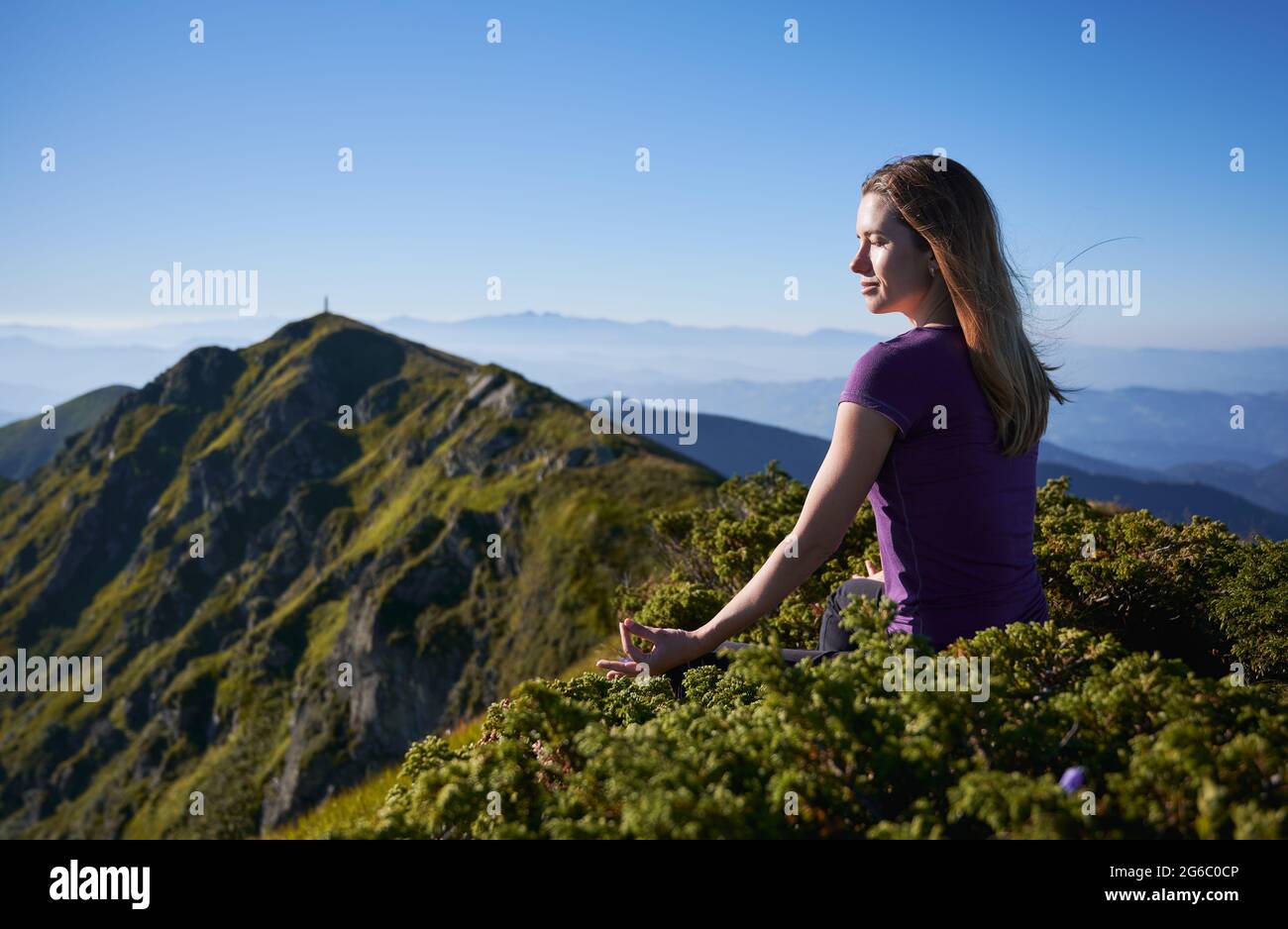 Bella giovane donna seduta su collina erbosa in posizione di loto e facendo mudra Gyan gesto mano mentre meditando all'aperto. Donna sportiva serena che fa meditazione yoga esercizio in montagna. Foto Stock