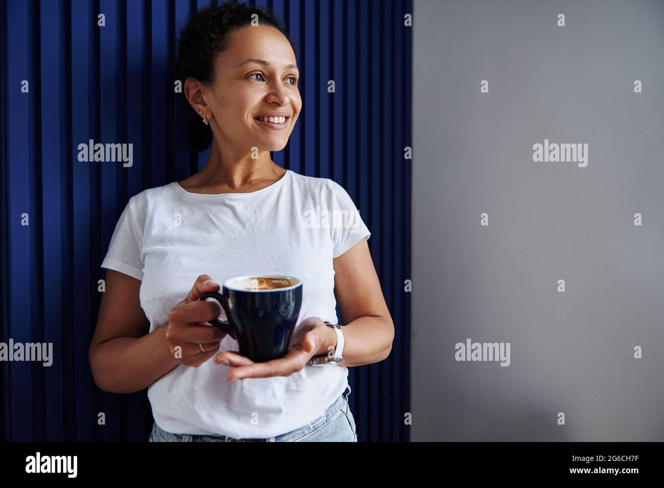 Donna matura con bruna sorridente con una tazza di caffè in mani appoggiata su una parete blu goffrata. Ritratto di bella donna con tazza di bevanda calda Foto Stock